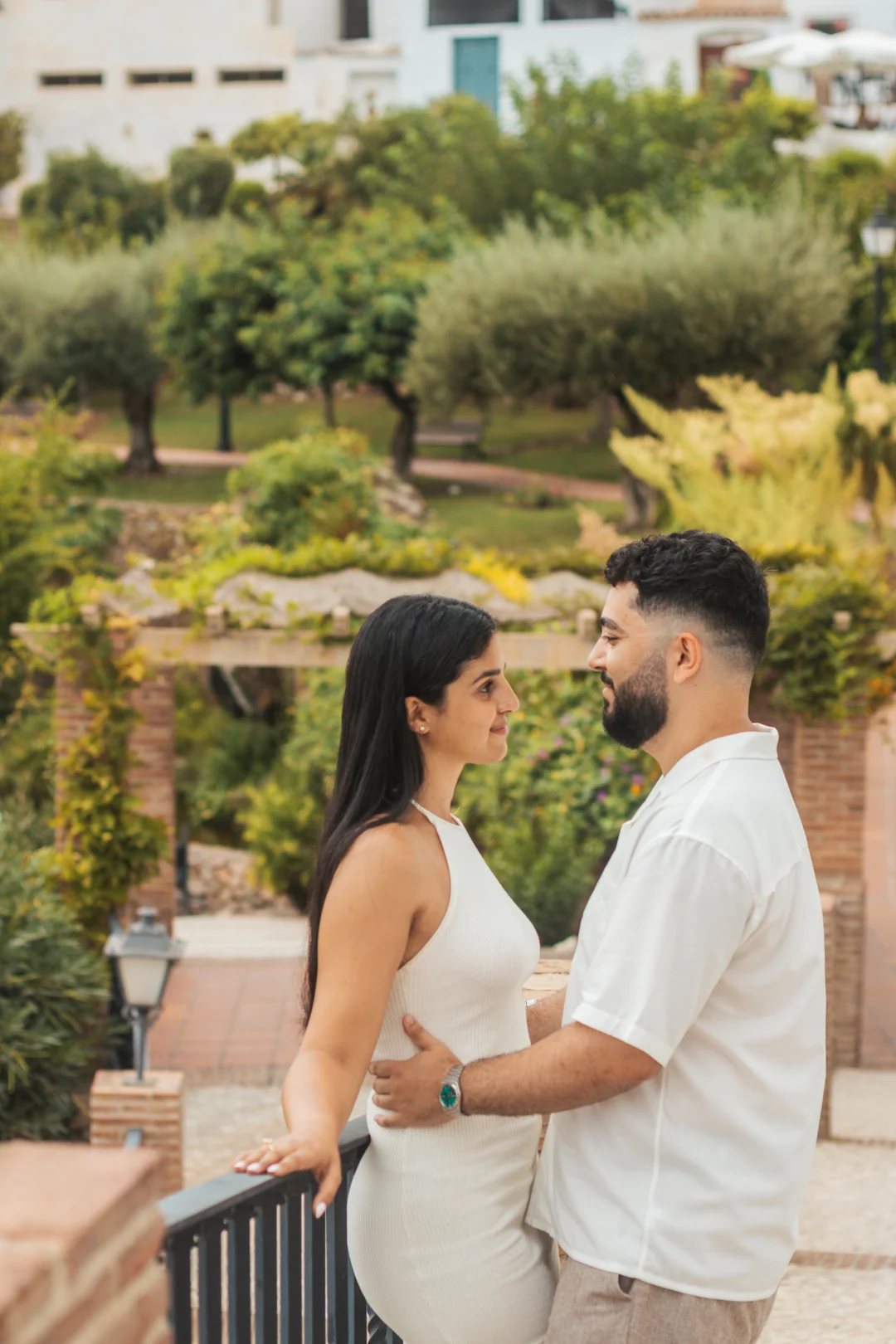 An intimate moment between a couple on a sun-drenched Mediterranean terrace, with a lush green garden backdrop typical of Andalusia's coastal landscape.