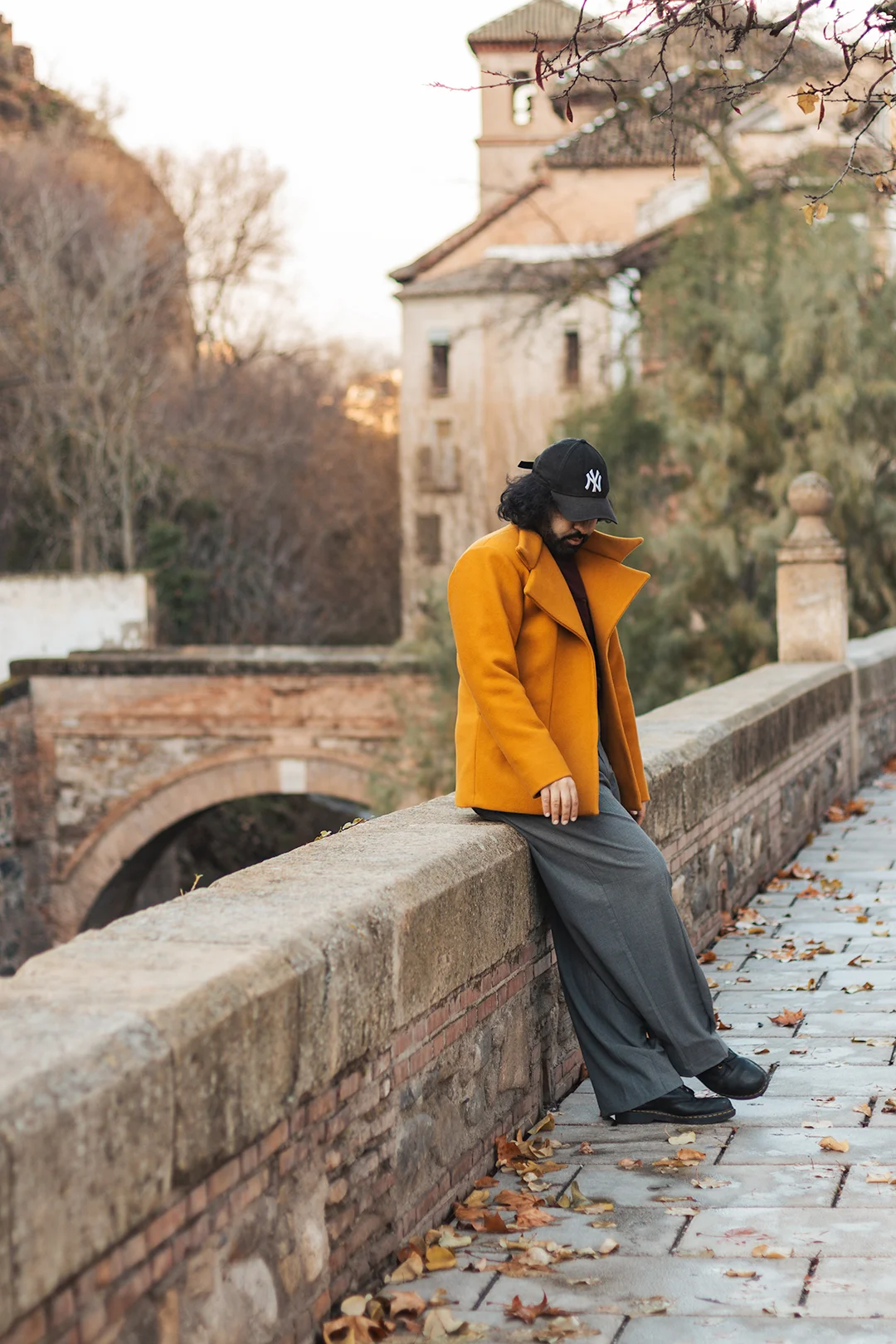 An individual portrait full of autumn mood — a young person leans on a centuries-old stone bridge in Granada's Río Darro area, golden leaves scattered on the cobblestones below. An individual portrait full of autumn mood — a young person leans on a centuries-old stone bridge in Granada's Río Darro area, golden leaves scattered on the cobblestones below.