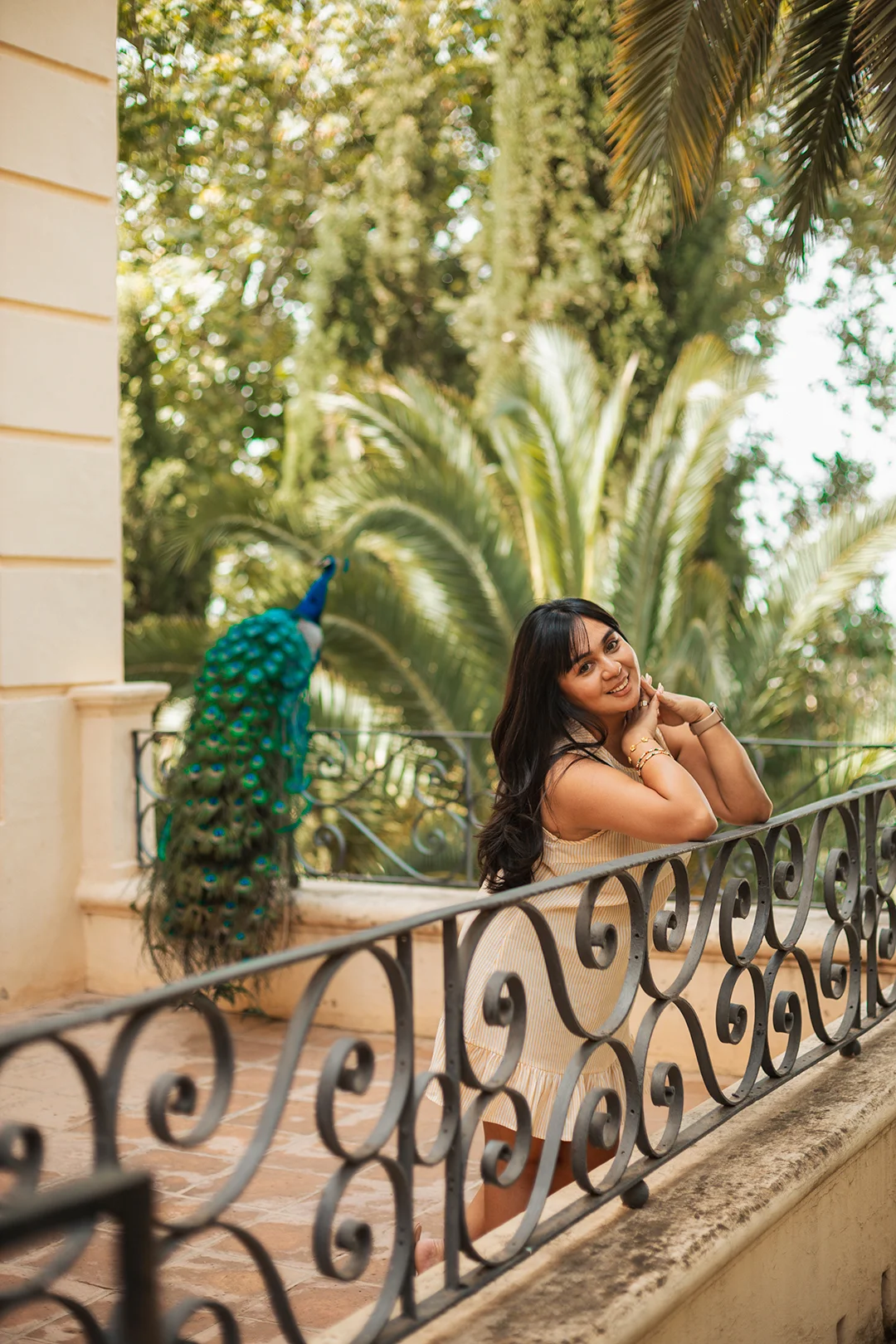 An enchanting individual portrait — a woman poses gracefully on a wrought-iron terrace railing with a magnificent peacock standing nearby, surrounded by a lush tropical garden in Andalusia. An enchanting individual portrait — a woman poses gracefully on a wrought-iron terrace railing with a magnificent peacock standing nearby, surrounded by a lush tropical garden in Andalusia.