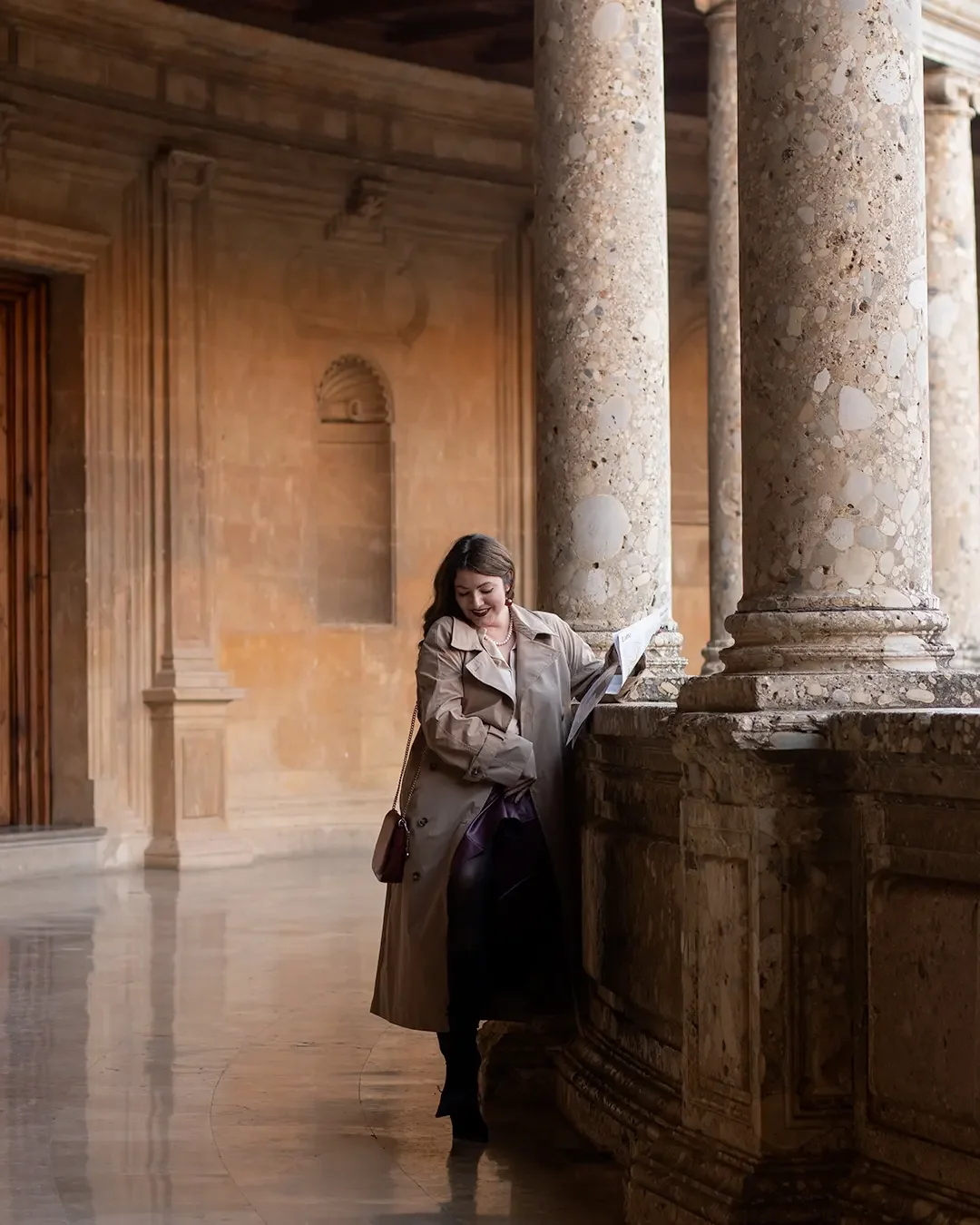 A poetic individual portrait — a woman in a camel trench coat reads quietly among the magnificent marble columns of the Palace of Charles V inside Granada's legendary Alhambra complex. A poetic individual portrait — a woman in a camel trench coat reads quietly among the magnificent marble columns of the Palace of Charles V inside Granada's legendary Alhambra complex.
