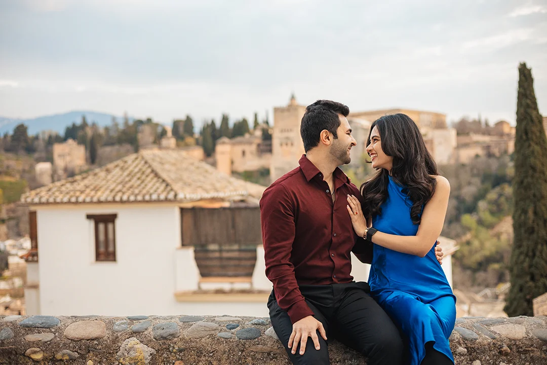 A romantic couple shares a smile while sitting on a stone ledge overlooking the historic Alhambra palace in Granada, showcasing professional photography in the Albaicín district. A romantic couple shares a smile while sitting on a stone ledge overlooking the historic Alhambra palace in Granada, showcasing professional photography in the Albaicín district.